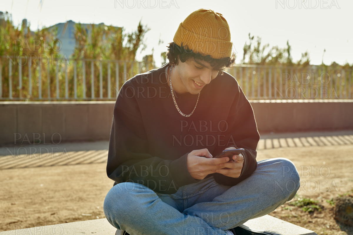 Smiling cross-legged guy using cellphone outdoors stock photo